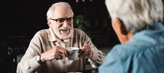 happy senior man in eyeglasses holding cup of coffee near blurred friend in cafe, banner