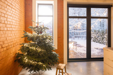 Cozy living room interior with Christmas tree in the corner. Concept of home comfort in winter. Windows overlooking a snowy garden, home with brick wall