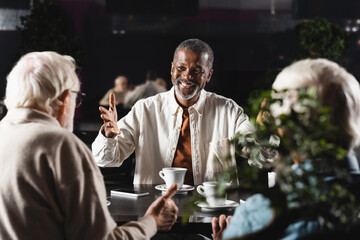 happy african american man pointing with hands at blurred senior friends in cafe