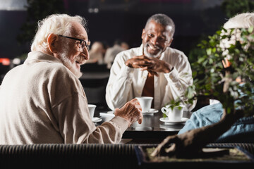 senior man in eyeglasses talking to interracial friends spending time in cafe