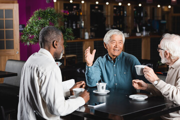 Fototapeta na wymiar smiling asian man gesturing while talking with senior multiethnic friends in cafe