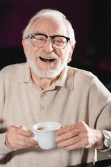 cheerful senior man in eyeglasses holding cup of coffee while laughing at camera