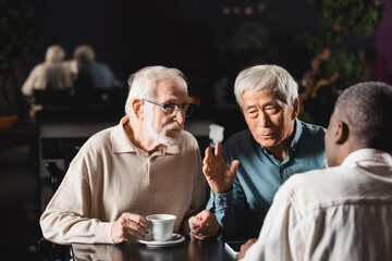 Fototapeta na wymiar elderly asian man talking and gesturing near multiethnic friends in cafe