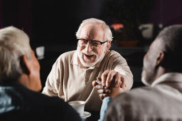 excited senior man in eyeglasses joining hands with blurred multiethnic friends in bar