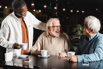 senior african american man with cup of coffee standing near cheerful interracial friends in cafe