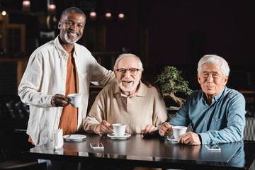 elderly multiethnic friends smiling at camera while drinking coffee in restaurant