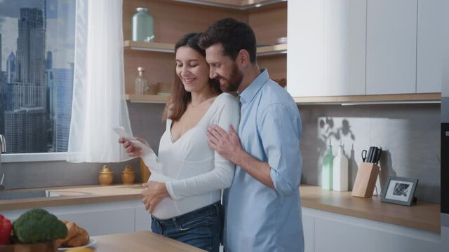 Caucasian cheerful couple of future affectionate parents pregnant wife and husband watching on ultrasound photograph in modern stylish cozy home kitchen.