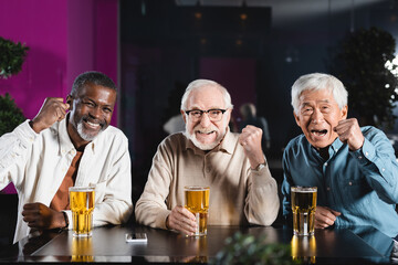 happy senior multiethnic friends showing win gesture while watching football championship in pub