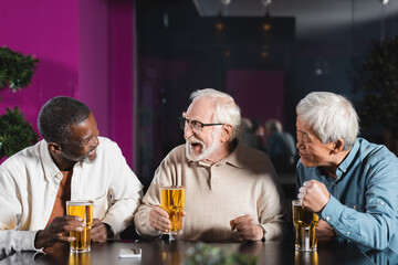 excited elderly multicultural friends talking in pub near glasses of beer