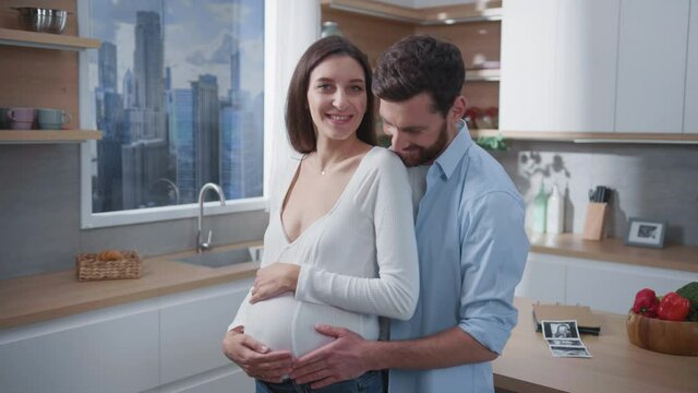 Husband with pretty pregnant young wife holding hands together embracing in the kitchen. Family life. Couple portrait. Affectionate future parents.