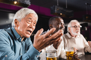 discouraged asian man pointing with hand while watching football match with smiling interracial friends in pub