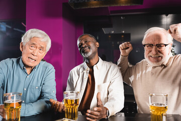 excited senior man showing success gesture while watching football match with upset interracial friends