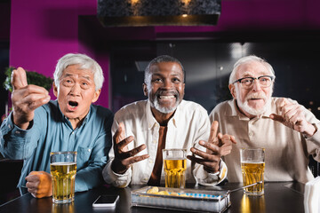 emotional senior multicultural friends watching football match near glasses of beer and chips in pub