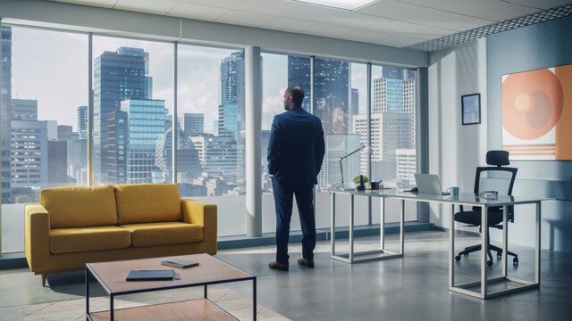 Thoughtful African-American Businessman in a Perfect Tailored Suit Standing in His Office Looking out of the Window on Big City. Successful CEO Planning Investment Strategy for e-Commerce Startup