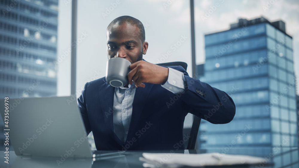 Poster portrait of black businessman in tailored suit working on laptop computer and drinking coffee in his