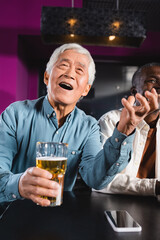 amazed asian man with glass of beer watching championship in sports bar near senior multiethnic friends