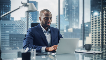 Fototapeta premium Successful Black Businessman in Tailored Suit Working on Laptop Computer on Top Floor Office Overlooking Big City. Professional CEO Managing Environmental, Social and Corporate Governance