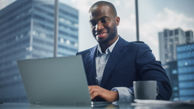 Portrait of Successful Black Businessman in Tailored Suit Working on Laptop Computer in His Big City Office. Digital Entrepreneur does Data Analysis, Cloud Computing for e-Commerce Strategy assessment