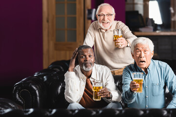 thrilled senior multicultural friends with glasses of beer watching football match in sports bar