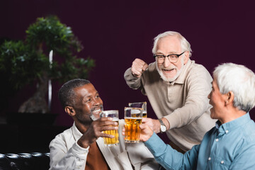 excited senior man showing win gesture while clinking glasses of beer with friends in pub