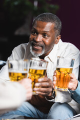 senior african american man smiling while clinking blurred beer glasses with friends