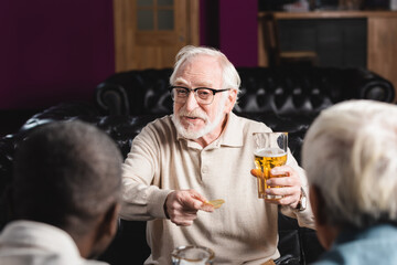 cheerful senior man holding chips and beer while talking to blurred interracial friends in pub