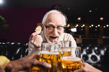 excited senior man holding chips while clinking beer glasses with blurred interracial friends