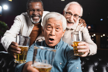 cheerful senior multicultural friends looking at camera while holding glasses of beer in pub
