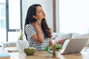 Tired business woman with neck pain looking uncomfortable while working with laptop in living room at home.