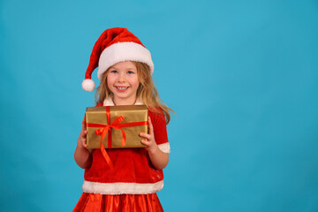 christmas little girl smiling with a gift on the background for the text in a santa costume and a cap. Happy Holidays and present a box with a bow. Copy space. Happy child for the new year.