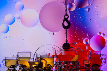 transparent glass teapot and cups with tea and drops, silhouette of lifting hook on colorful background 