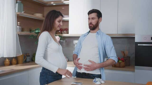 Family life. Fun scene of happy woman pregnant with child laughing of her husband pretending to be pregnant. Young parents. Home kitchen interior.