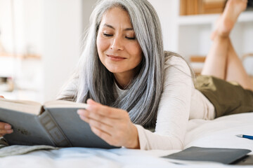 Grey asian woman smiling and reading book while lying on bed