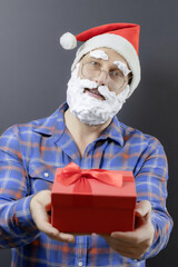 Santa in a plaid shirt with a white foam beard is holding a red gift box. Focus on the face. Merry Christmas to you