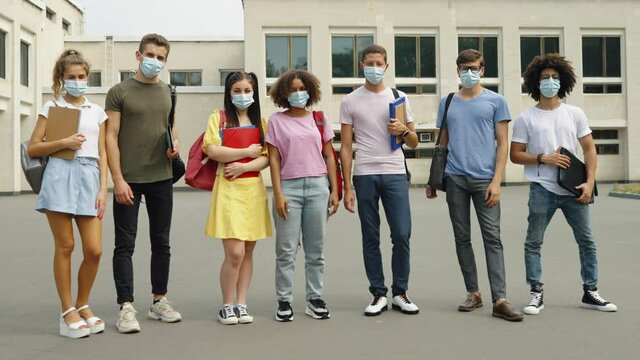 Group of students wearing protective masks standing with backpacks and books against college building, looking at camera. Tracking shot of diverse young people during pandemic. Concept of education