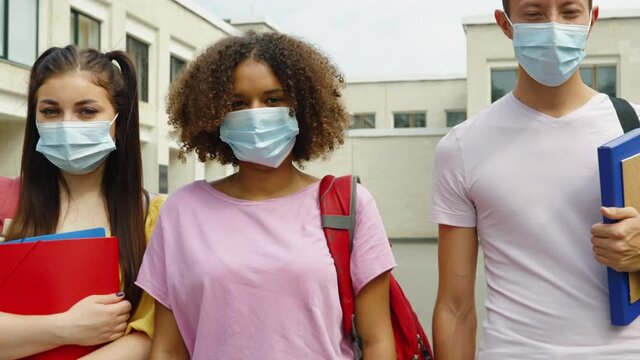 Students wearing protective masks standing in line against college building with backpacks and books, looking at camera, camera zoom out. Concept of education during pandemic