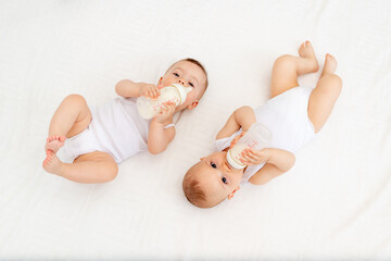 twin babies boy and girl with a bottle of milk on a white bed at home, baby food concept, place for text - Powered by Adobe