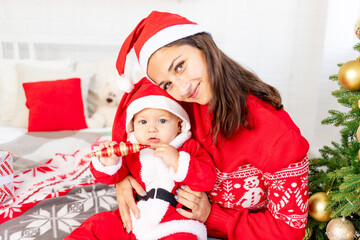 New Year or Christmas, a young mother with a baby on the bed at home by the Christmas tree in a Santa Claus costume hugging and waiting for the holiday smiling