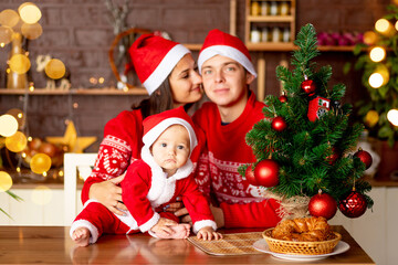 Fototapeta na wymiar New Year or Christmas, close-up of a baby in a red sweater and Santa Claus hat in the kitchen, a happy young family mom, dad and baby at the Christmas tree smiling, hugging
