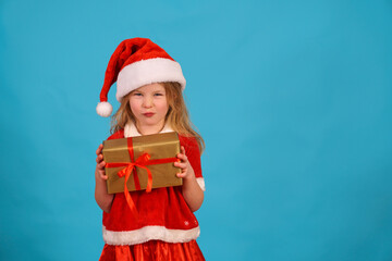 Funny Christmas child. girl in santa claus cap is holding gift and grimacing. Play with facial expressions. Sly look of satisfied girl with gift. Mischievous and naughty with gift on blue background.