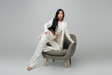 Fototapeta na wymiar full length of young asian woman leaning on armchair while posing with hand in pocket on grey