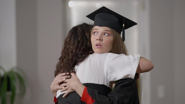 Young female graduate in gown and hat hugging woman with dissatisfied irritated facial expression. Caucasian hypocritical woman embracing mother on graduation day. Egoism concept