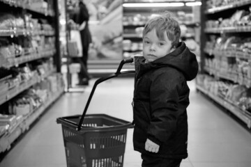 A little boy got lost among the shelves with goods in the store. A kid with a shopping basket in...