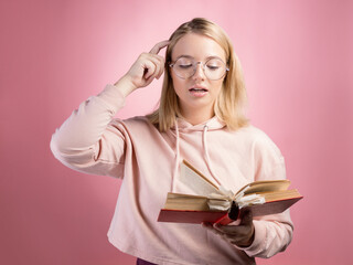 Think. A young female student with a book in her hands is studying the topic. A young cute blonde in a pink hoodie and glasses, a gesture of thoughtfulness, a finger on her forehead.