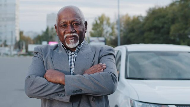 Old stately handsome african american man closeup stands near white car elderly person folded arms across chest serious face businessman ethnic male gray beard in city outdoors retiree posing thinks