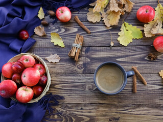 Latte mug, ripe red apples basket, blue plaid, scarf, fall oak leaves, spices, cinnamon sticks top view on dark wooden table. Autumn hot drink, food concept