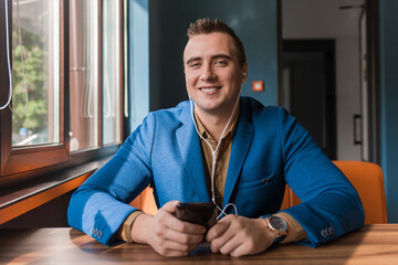 Businessman, a stylish, handsome smiling guy of Caucasian appearance portrait in a blue jacket spends time in a smartphone or mobile phone sitting at a table in the cafe