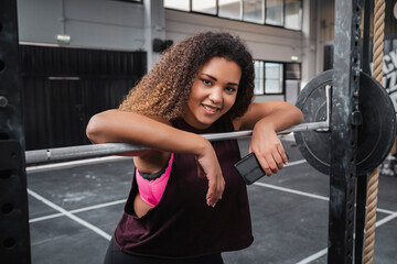 Smiling female athlete with mobile phone leaning on barbell at gym