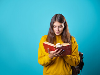 A student with a book in her hands, reading a textbook and preparing for a new lesson. A young brunette in a yellow hoodie and with a backpack, leafing through a book.