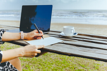 Woman working on laptop by the beach with hand writing in paper notebook.